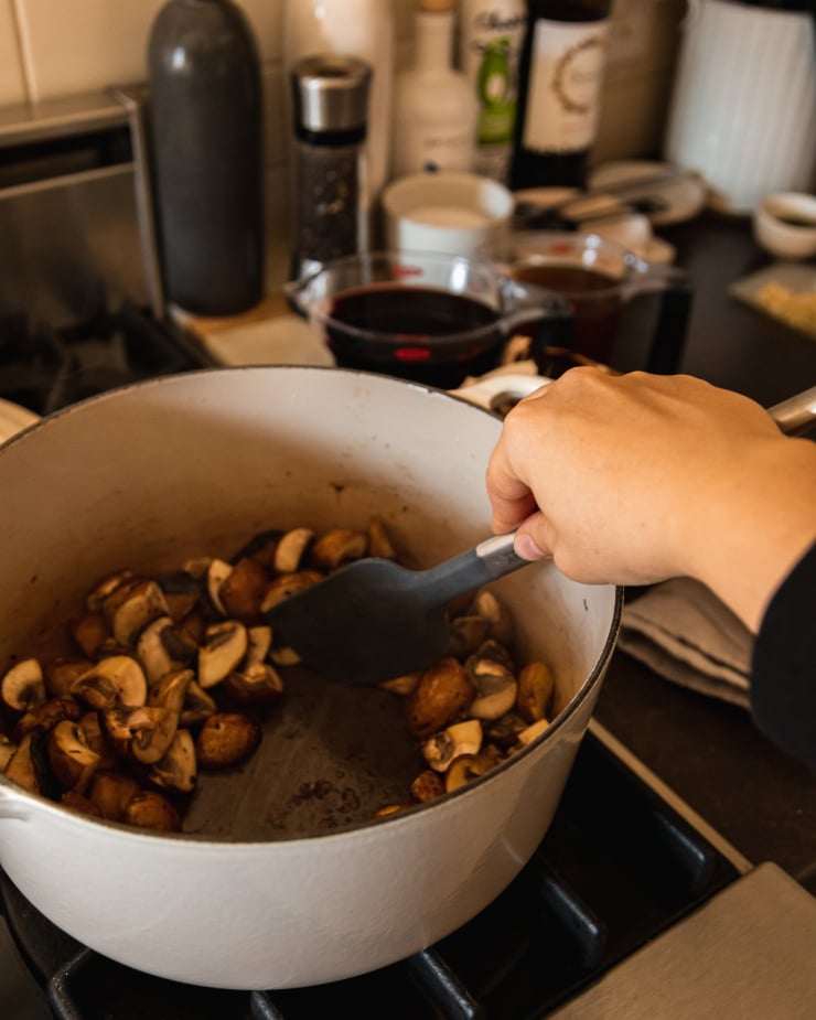 Image shows. hand using a spatula to stir sautรฉed mushrooms in a big off-white pot over a gas stove.