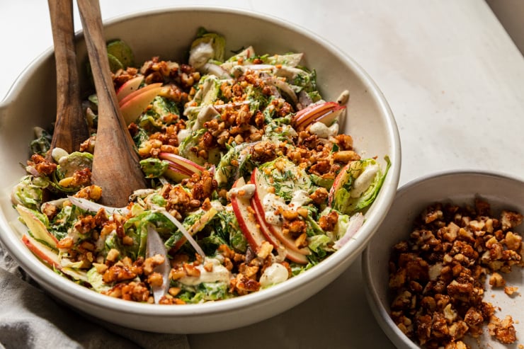 A 3/4 angle shot of a shaved brussels sprout salad with sliced apples, toasted walnuts, and sliced red onion on top. A creamy white dressing is drizzled over the salad as well.