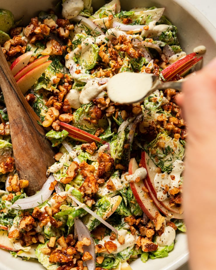 A hand is using a spoon to drizzle creamy dressing over a Fall salad with Brussels sprouts, apples, chopped candied walnuts, and apples.