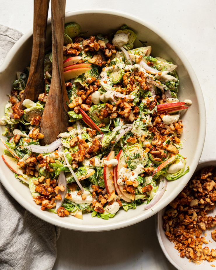 An overhead shot of a shaved brussels sprout salad with sliced apples, toasted walnuts, and sliced red onion on top. A creamy white dressing is drizzled over the salad as well.