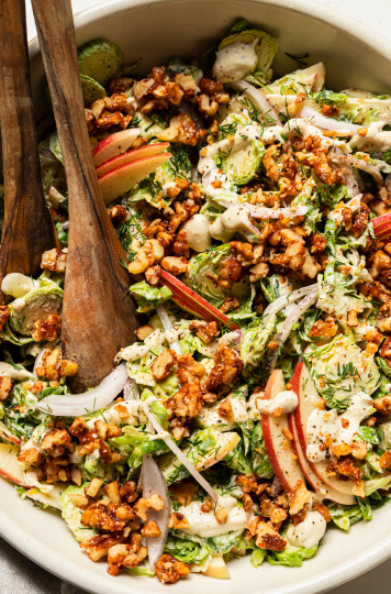 An up close, overhead shot of a shaved brussels sprout salad with sliced apples, toasted walnuts, and sliced red onion on top. A creamy white dressing is drizzled over the salad as well.