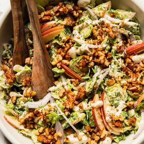 An up close, overhead shot of a shaved brussels sprout salad with sliced apples, toasted walnuts, and sliced red onion on top. A creamy white dressing is drizzled over the salad as well.