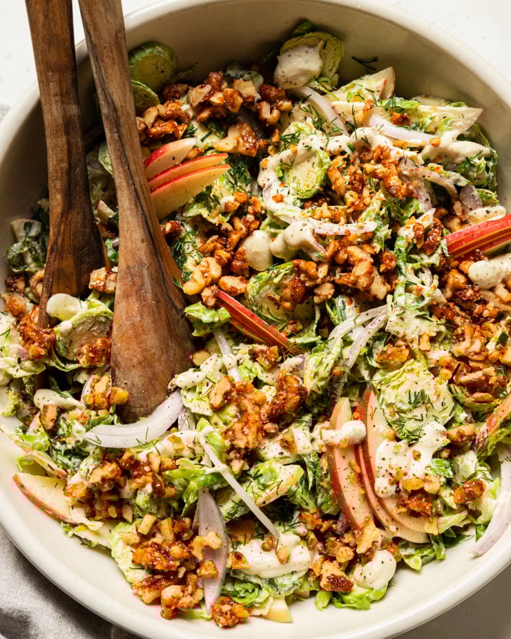 An up close, overhead shot of a shaved brussels sprout salad with sliced apples, toasted walnuts, and sliced red onion on top. A creamy white dressing is drizzled over the salad as well.