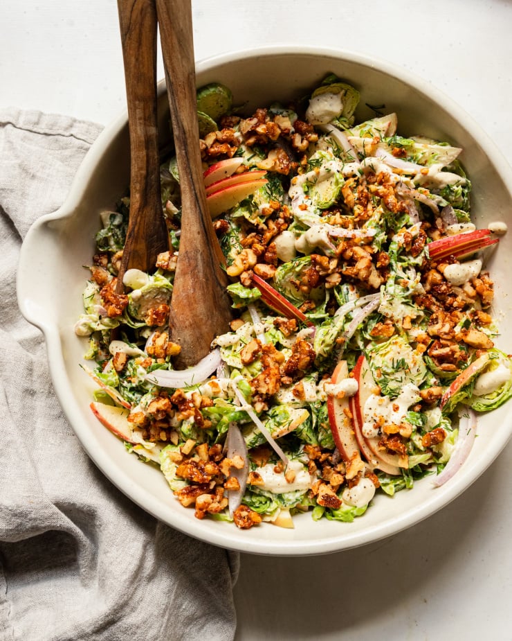 An overhead shot of a shaved brussels sprout salad with sliced apples, toasted walnuts, and sliced red onion on top. A creamy white dressing is drizzled over the salad as well.