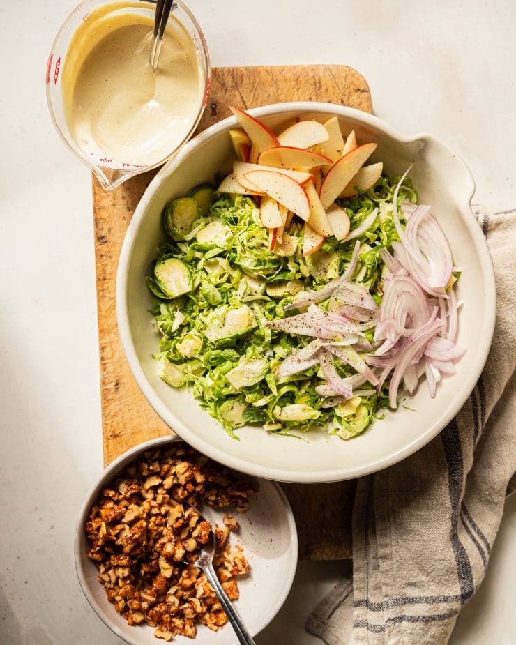An overhead shot of components for a brussels sprout salad: a large bowl with shaved brussels sprouts, red onion & apples, a measuring cup with creamy dressing, and a small bowl with chopped candied walnuts.