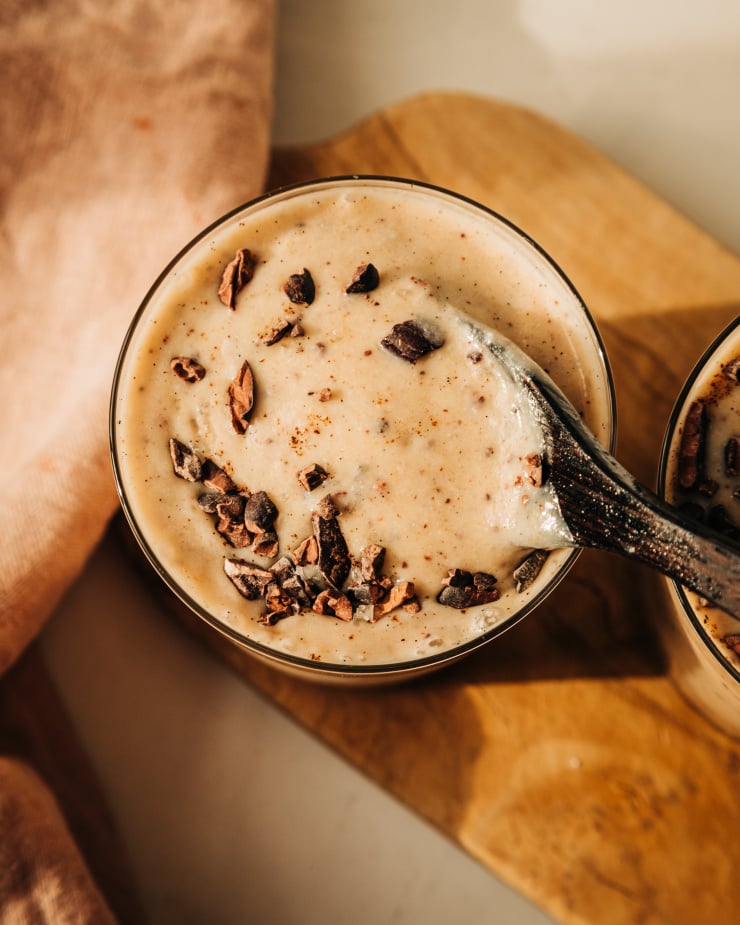 An overhead and up close shot of a beige smoothie being stirred up with a dark wooden spoon. The smoothie is topped with cacao nibs.