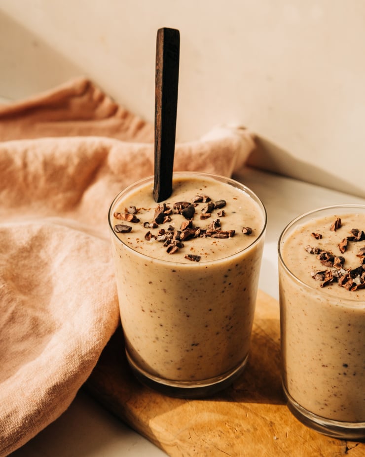 A 3/4 angle shot of a creamy beige smoothie with dark brown flecks. A wooden spoon is sticking out of the glass.