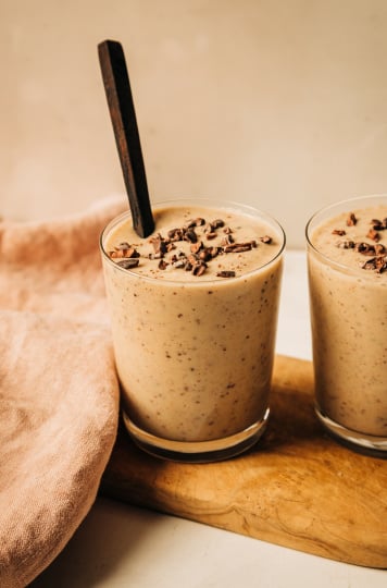 A head on shot of a creamy, beige vanilla chip smoothie in two clear glasses. The smoothie is garnished with cacao nibs and there is a dark wooden spoon sticking out of the one glass.