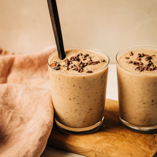 A head on shot of a creamy, beige vanilla chip smoothie in two clear glasses. The smoothie is garnished with cacao nibs and there is a dark wooden spoon sticking out of the one glass.