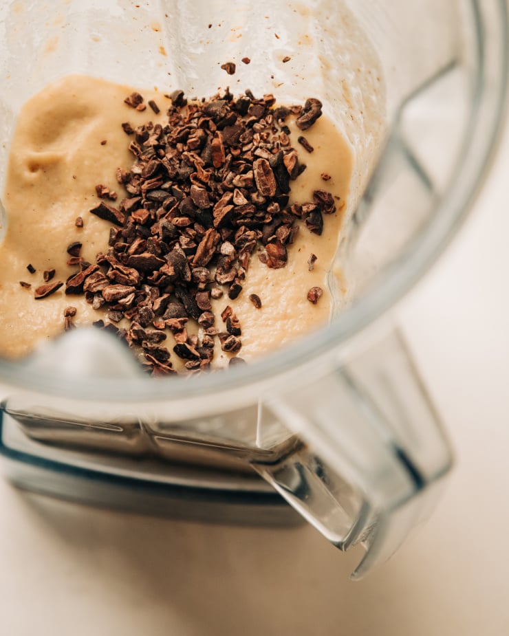 An overhead shot of cacao nibs on top of a blended beige concoction inside of a blender pitcher.