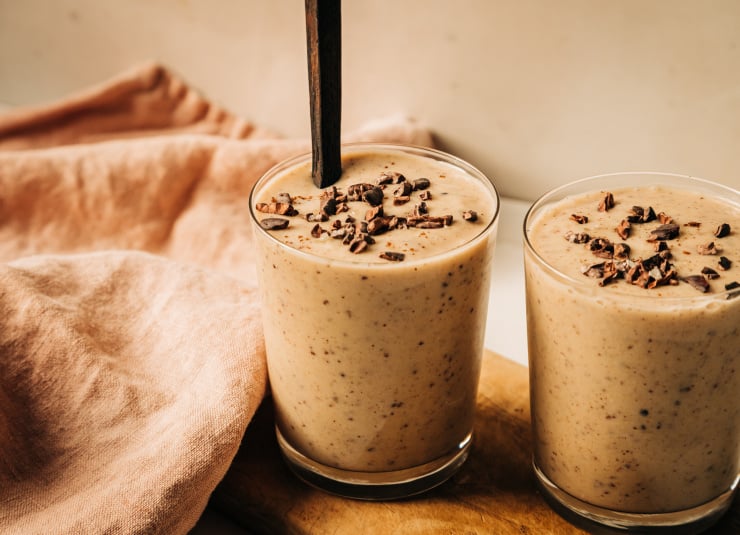 A head on shot of a creamy beige smoothie flecked with brown bits in two clear glasses.