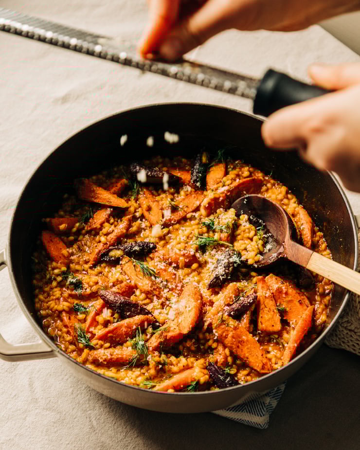 A 3/4 angle image shows a hand grating a walnut into shavings over a barley and roasted carrot risotto in a braiser-style pot.