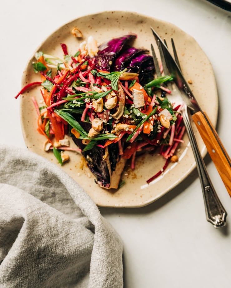 An overhead shot of a roasted red cabbage wedge on a beige speckled plate. The wedge is topped with a root vegetable and herb slaw. The portion is topped with chopped toasted cashews, flaked coconut, and sesame seeds.