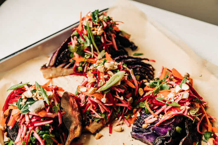 A 3/4 angle shot of roasted red cabbage wedges on a parchment-lined white platter. The wedges are topped with a root vegetable and herb slaw. Each wedge/slaw portion is topped with chopped toasted cashews, flaked coconut, and sesame seeds.