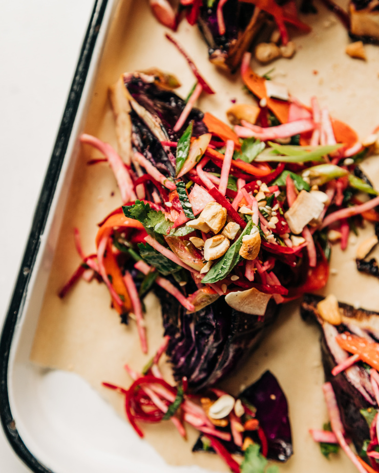 An overhead shot of roasted red cabbage wedges on a parchment-lined white platter. The wedges are topped with a root vegetable and herb slaw. Each wedge/slaw portion is topped with chopped toasted cashews, flaked coconut, and sesame seeds.
