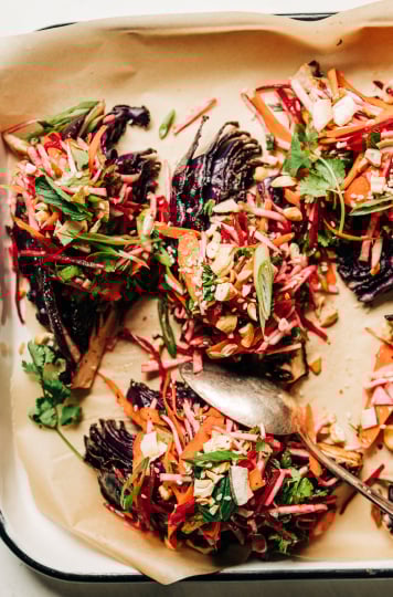 An overhead shot of roasted red cabbage wedges on a parchment-lined white platter. The wedges are topped with a root vegetable and herb slaw. Each wedge/slaw portion is topped with chopped toasted cashews, flaked coconut, and sesame seeds.