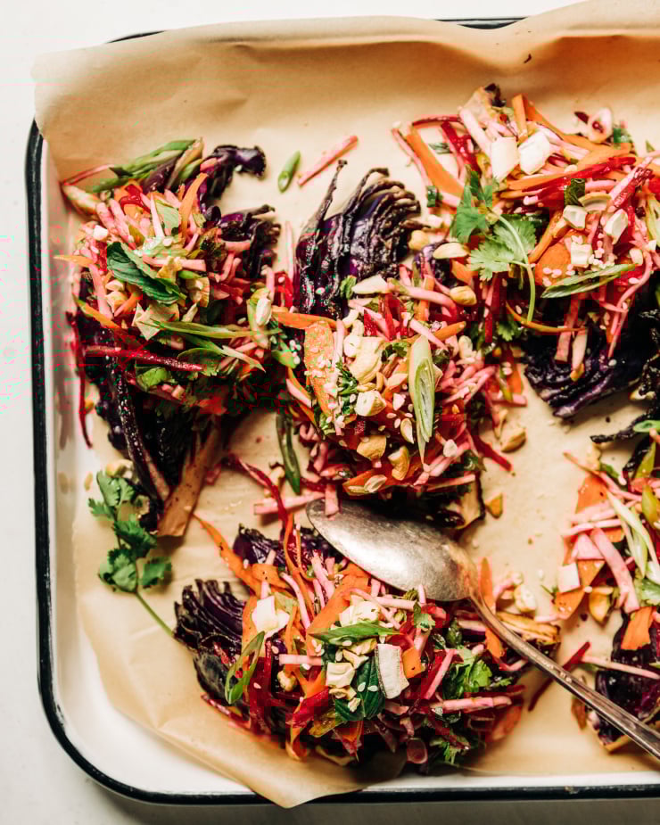 An overhead shot of roasted red cabbage wedges on a parchment-lined white platter. The wedges are topped with a root vegetable and herb slaw. Each wedge/slaw portion is topped with chopped toasted cashews, flaked coconut, and sesame seeds.