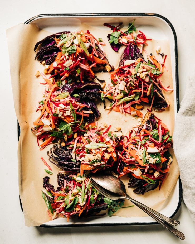 An overhead shot of roasted red cabbage wedges on a parchment-lined white platter. The wedges are topped with a root vegetable and herb slaw. Each wedge/slaw portion is topped with chopped toasted cashews, flaked coconut, and sesame seeds.
