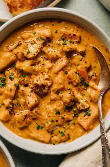 An overhead shot of a creamy orange vegetable stew in a white bowl with an antique spoon sticking out. The stew is a cauliflower and sweet potato chowder and is garnished with finely minced chives.