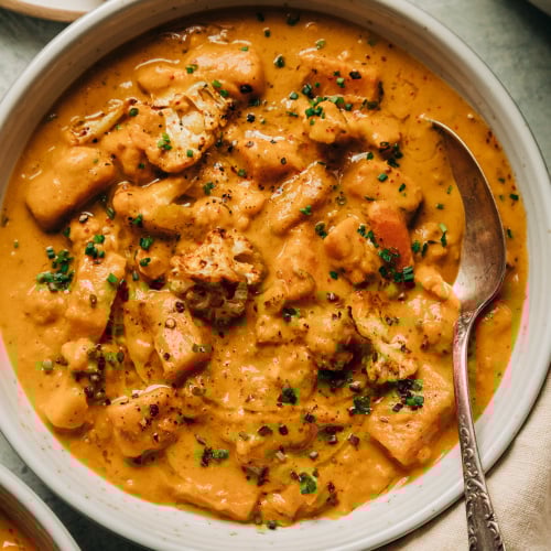 An overhead shot of a creamy orange vegetable stew in a white bowl with an antique spoon sticking out. The stew is a cauliflower and sweet potato chowder and is garnished with finely minced chives.