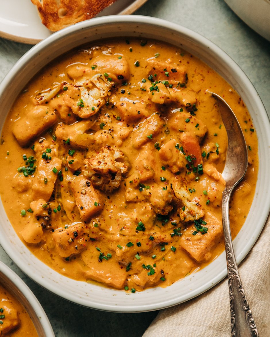 An overhead shot of a creamy orange vegetable stew in a white bowl with an antique spoon sticking out. The stew is a cauliflower and sweet potato chowder and is garnished with finely minced chives.