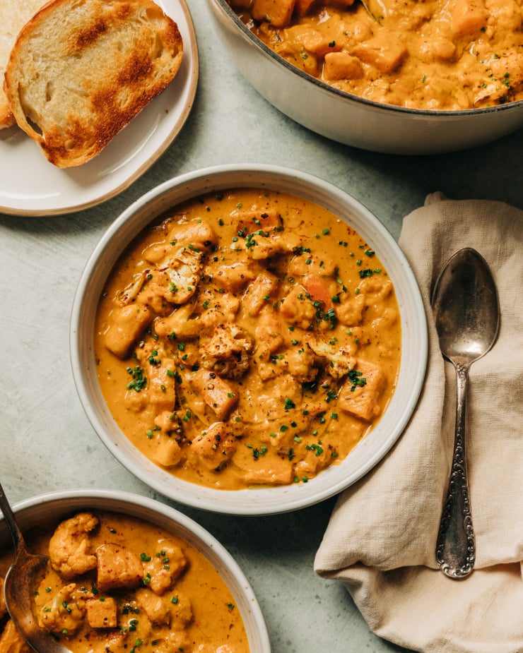 An overhead shot of a creamy orange vegetable stew in a white bowl with an antique spoon sticking out. The stew is a cauliflower and sweet potato chowder and is garnished with finely minced chives. A plate with toasted bread slices is seen nearby.