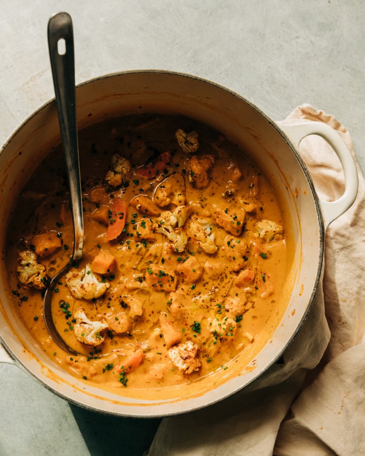 An overhead shot of a creamy orange stew with sweet potatoes and cauliflower in a white, Dutch oven-style pot. The stew is garnished with chopped chives.