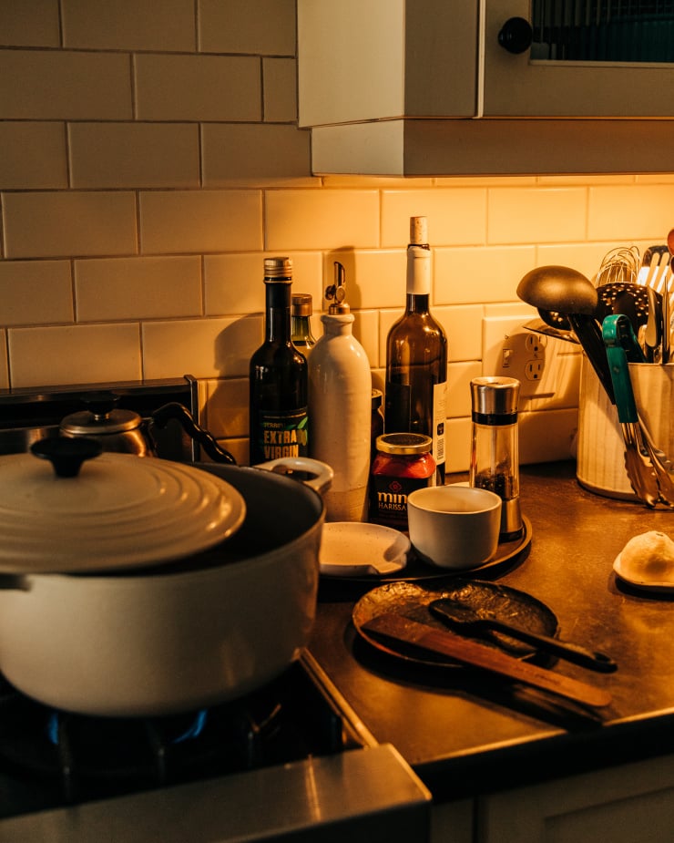 A dark kitchen scene shows a countertop and a gas stove with a Dutch oven-style pot on top. Against the subway tile background, we see bottles of cooking oil, a pepper grinder, and a carafe of salt. There is also a vase with a variety of cooking utensils sticking out.