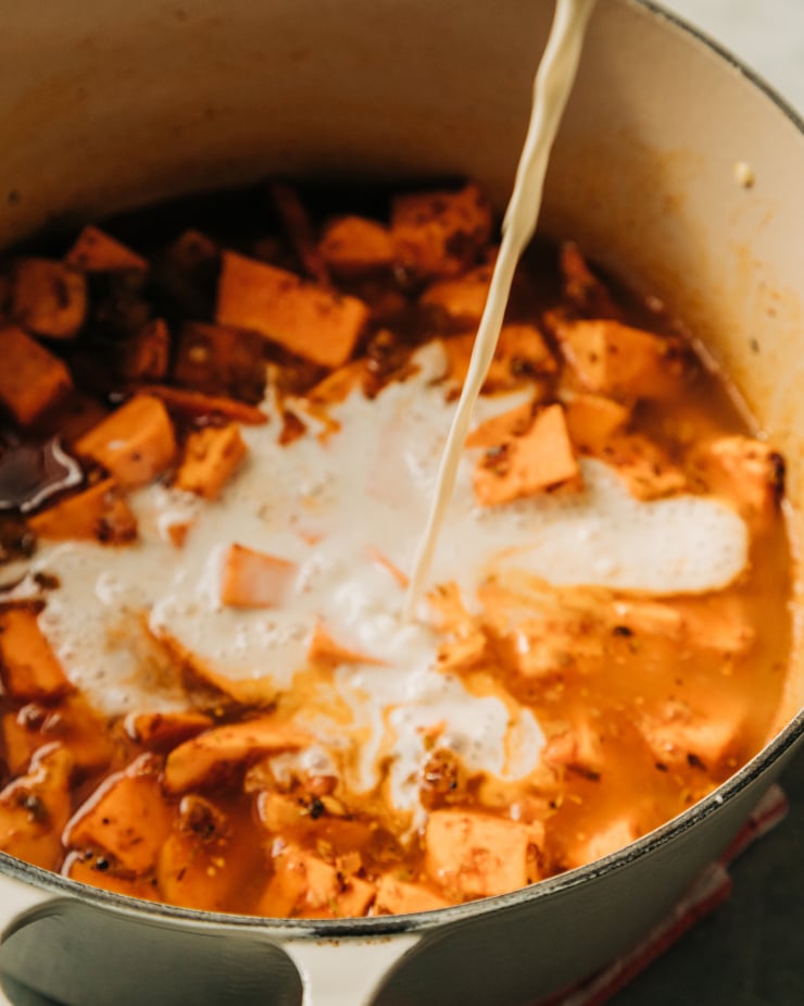 An up close shot of a non-dairy "cream" being poured into a pot with a soup-y sweet potato mixture inside.