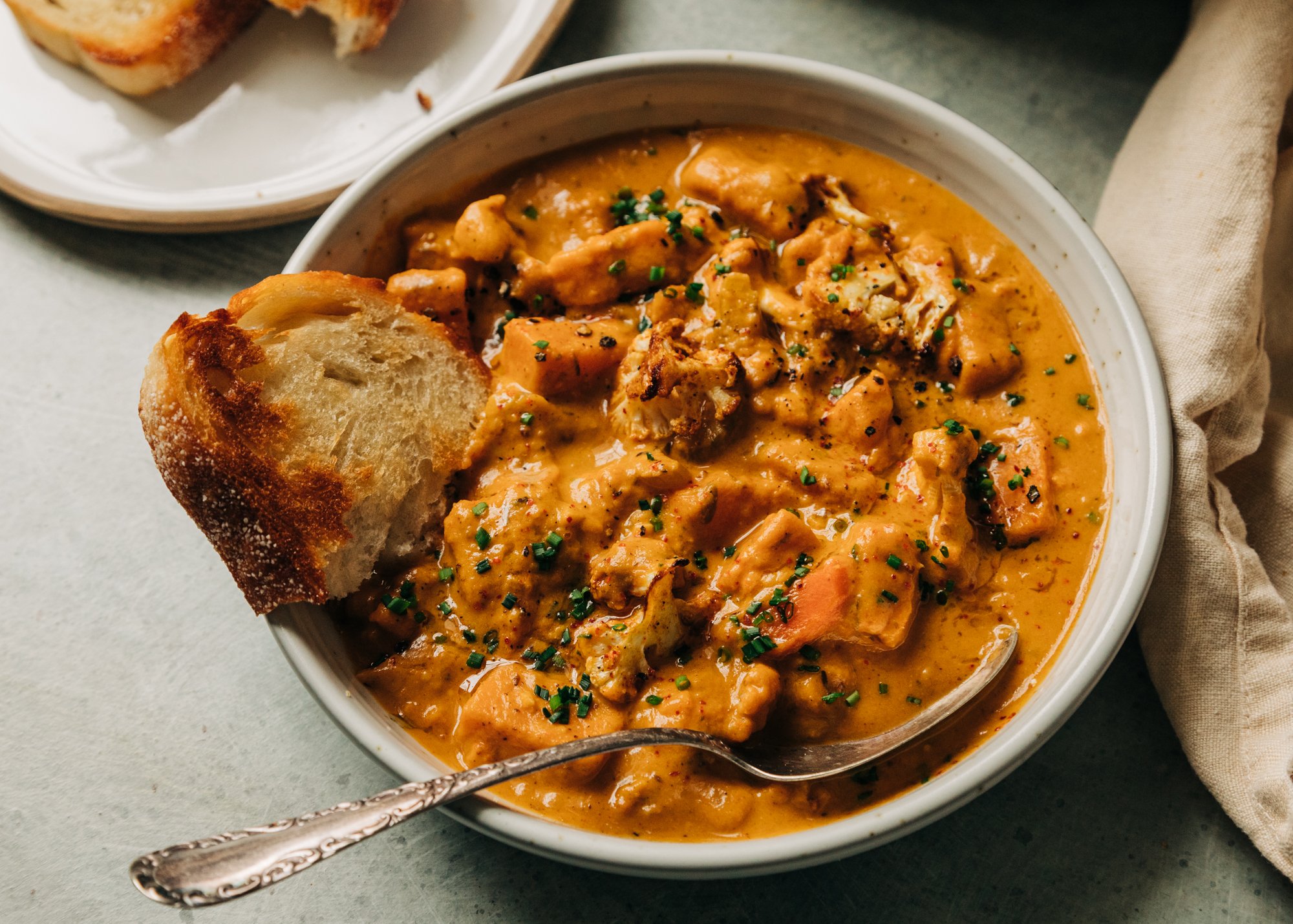 An 3/4 angle shot of a creamy orange vegetable stew in a white bowl with an antique spoon sticking out. The stew is a cauliflower and sweet potato chowder and is garnished with finely minced chives.