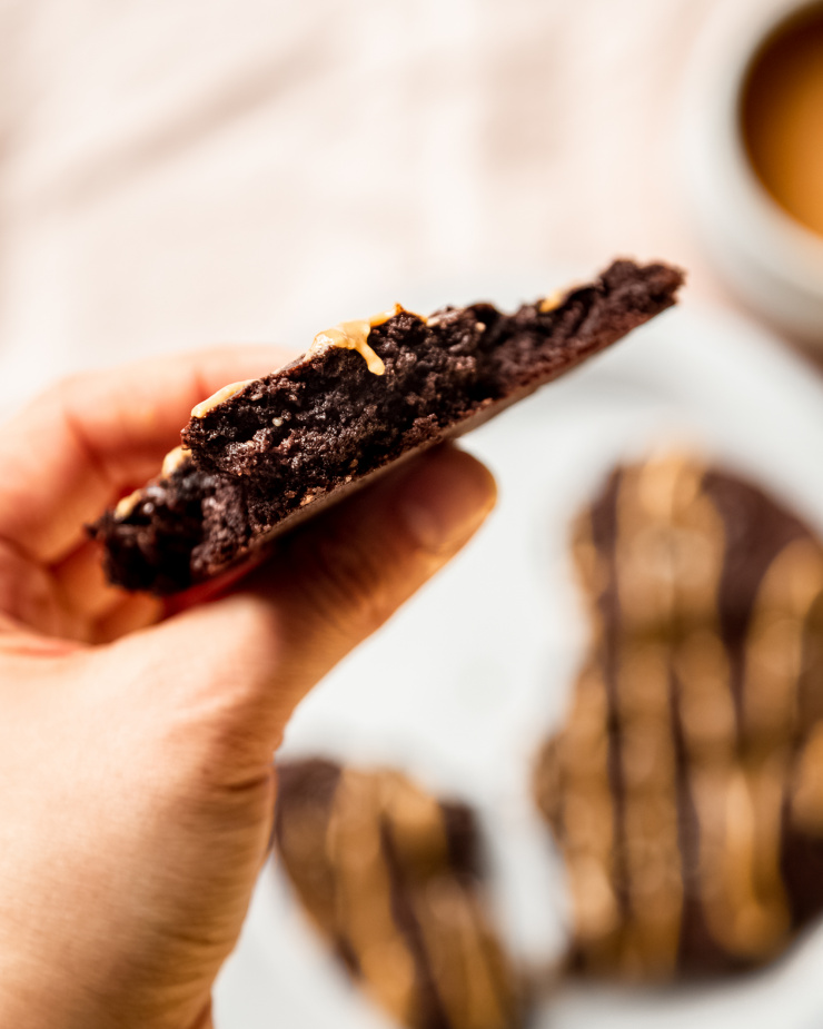 An overhead shot shows a hand holding half of a vegan Double Chocolate Cookie with a drip of peanut butter running down the middle.