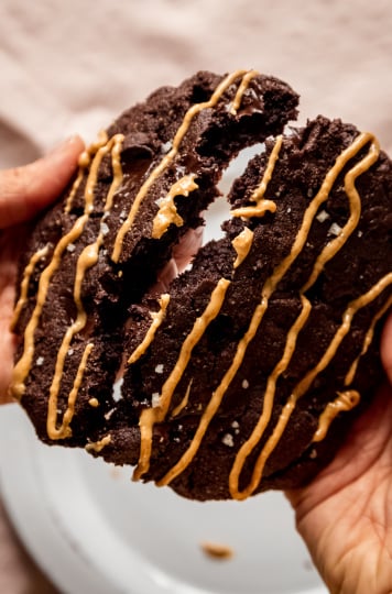 An overhead image shows a pair of hands splitting a jumbo double chocolate cookie drizzled with peanut butter in half.