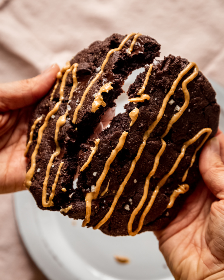 An overhead image shows a pair of hands splitting a jumbo double chocolate cookie drizzled with peanut butter in half.