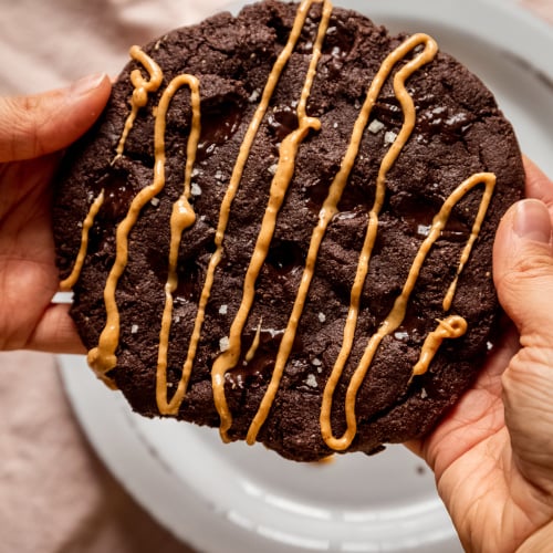 An overhead image shows a set of hands holding a jumbo double chocolate cookie drizzled with peanut butter.