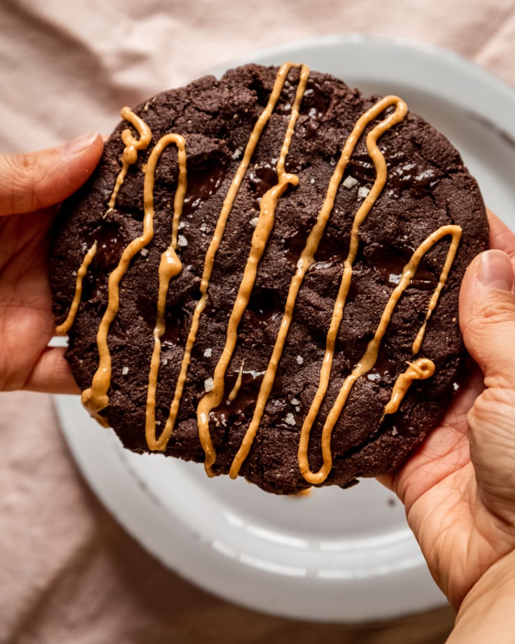 An overhead image shows a set of hands holding a jumbo double chocolate cookie drizzled with peanut butter.
