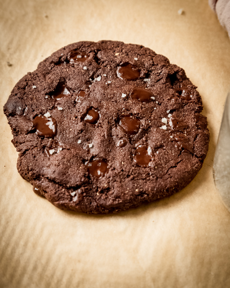 A slight 3/4 angle shot of a large double chocolate cookie garnished with flaky salt.