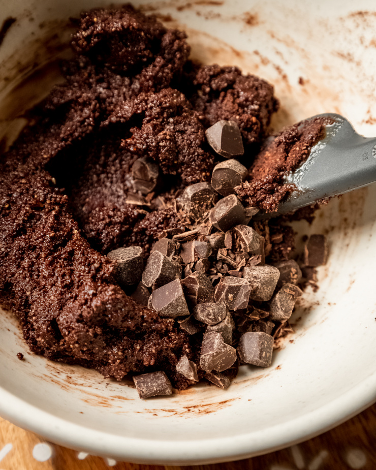 A 3/4 image shows chocolate chunks being stirred into a chocolate dough in a beige bowl with a spatula.