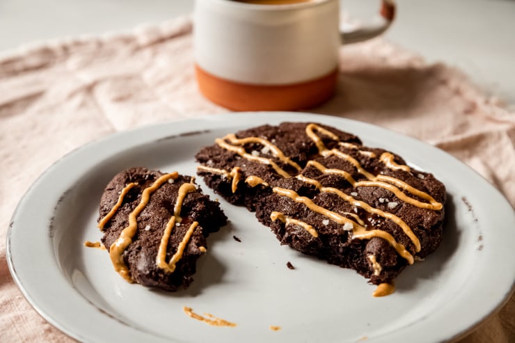A 3/4 angle image shows a large double chocolate cookie drizzled in peanut butter that has been split into pieces on a white plate.