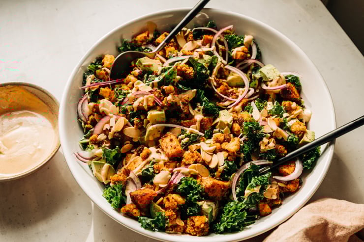 A 3/4 angle shot of a kale salad with creamy dressing on top, crispy bits of tofu, chunks of roasted sweet potato, slices of red onion, and cubes of avocado. The salad is in a white bowl.