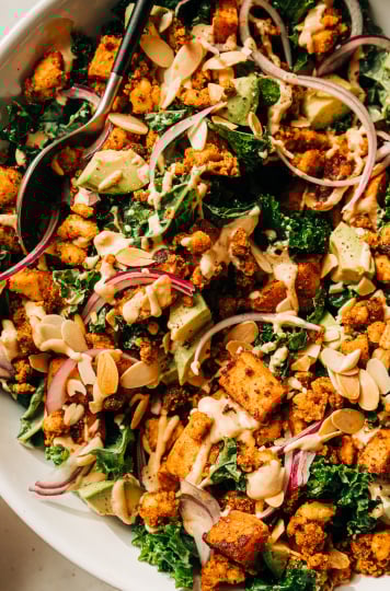 An overhead, up close shot of a kale salad with creamy dressing on top, crispy bits of tofu, chunks of roasted sweet potato, slices of red onion, and cubes of avocado. The salad is in a white bowl.