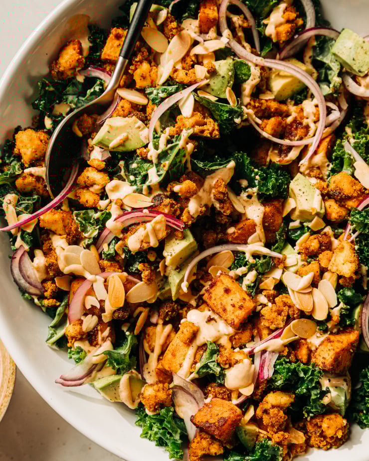 An overhead, up close shot of a kale salad with creamy dressing on top, crispy bits of tofu, chunks of roasted sweet potato, slices of red onion, and cubes of avocado. The salad is in a white bowl.