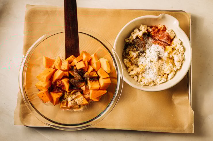 An overhead shot of two bowls on a parchment lined baking sheet. In one bowl is cubed sweet potatoes and spices. In the other is crumbled firm tofu and spices.