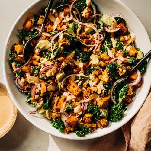 An overhead shot of a kale salad with creamy dressing on top, crispy bits of tofu, chunks of roasted sweet potato, slices of red onion, and cubes of avocado. The salad is in a white bowl.