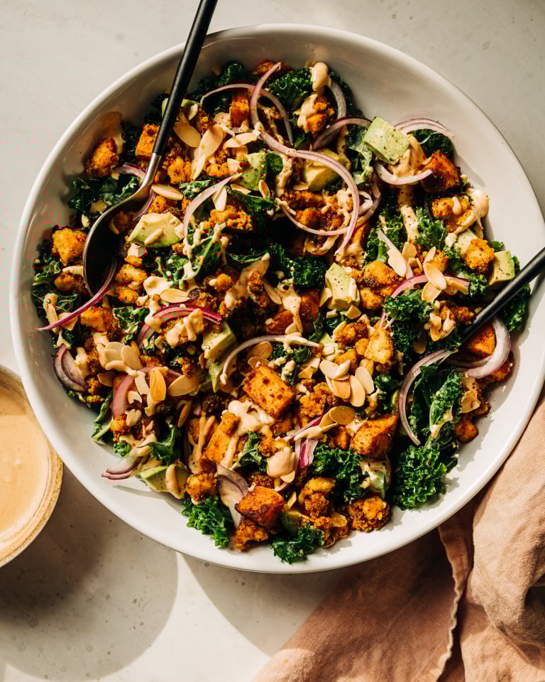 An overhead shot of a kale salad with creamy dressing on top, crispy bits of tofu, chunks of roasted sweet potato, slices of red onion, and cubes of avocado. The salad is in a white bowl.