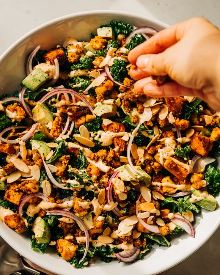 A hand is seen sprinkling crispy little bits of tofu over the top of a kale salad in a white bowl.