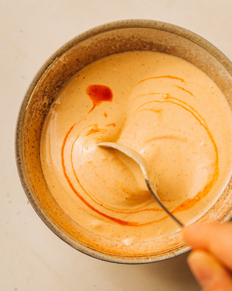 An overhead shot of a hand using a spoon to stir up a creamy, light orange sauce in a grey bowl.