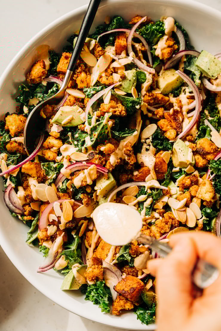 An overhead, up close shot of a kale salad with a hand using a spoon to drizzle a creamy dressing on top. The salad also features crispy bits of tofu, chunks of roasted sweet potato, slices of red onion, and cubes of avocado. The salad is in a white bowl.