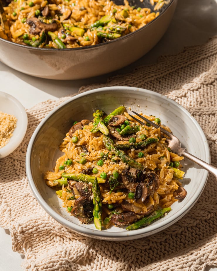 A 3/4 angle shot of a bowl of creamy orzo with spring vegetables. A pot of the orzo can be seen in the background.