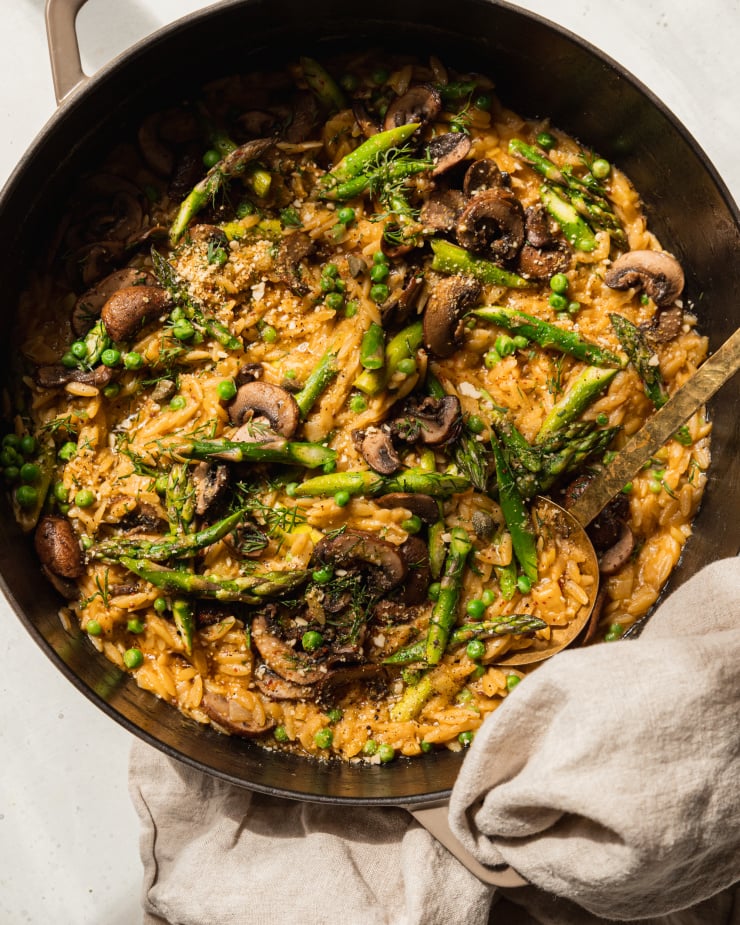 An overhead shot of creamy mushroom asparagus orzo with peas and bits of fresh dill on top. The orzo is in a braiser-style pot with a gold serving utensil sticking out. There is a beige napkin wrapped around the pot handle.