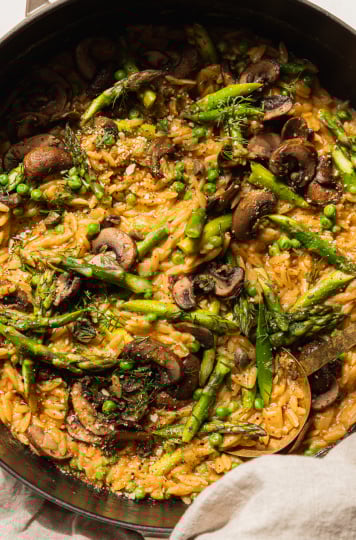 An overhead shot of creamy mushroom asparagus orzo with peas and bits of fresh dill on top. The orzo is in a braiser-style pot with a gold serving utensil sticking out. There is a beige napkin wrapped around the pot handle.
